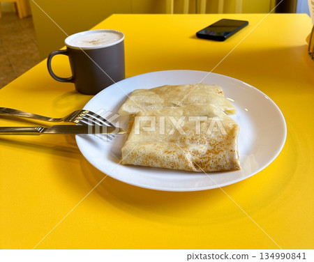 Crepes on white plate with coffee cup on yellow table. Simple breakfast, minimal food styling, cozy cafe mood and everyday comfort 134990841