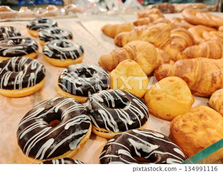 Chocolate Donuts Croissants Display. Assortment of chocolate-glazed donuts with white icing drizzle arranged alongside fresh buttery croissants on bakery counter. 134991116