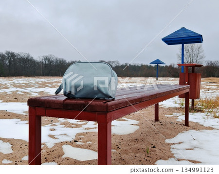 Placing cosmetic bag on wooden bench in snowy outdoor park area. Leaving small toiletry pouch on wet red bench near blue umbrella structures during overcast winter day 134991123