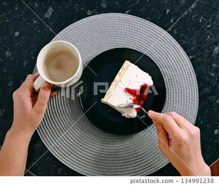 Eating Cheesecake Coffee Moment. Hands holding coffee mug and fork while eating piece of cheesecake with berry sauce on black plate viewed from above on table. Eating Cheesecake Coffee Moment. Hands holding coffee mug and fork while eating piece of cheesecake with berry sauce on black plate viewed from above on table. 134991238