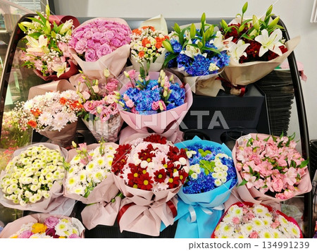 Flower Bouquets Shop Display. Multiple wrapped flower bouquets with roses, lilies, chrysanthemums and dyed flowers arranged on refrigerated stand in florist shop. 134991239