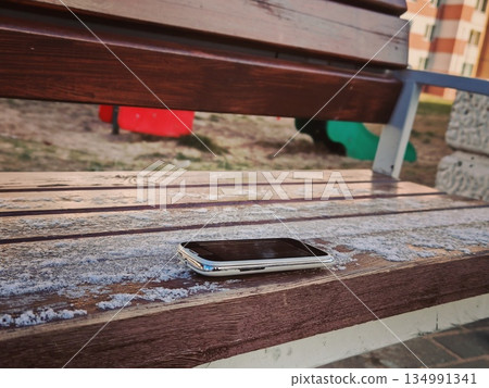 Forgotten Smartphone on Bench. Old mobile phone left lying on wooden park bench covered with light frost in outdoor playground area. Forgotten Smartphone on Bench. Old mobile phone left lying on wooden park bench covered with light frost in outdoor playground area. 134991341