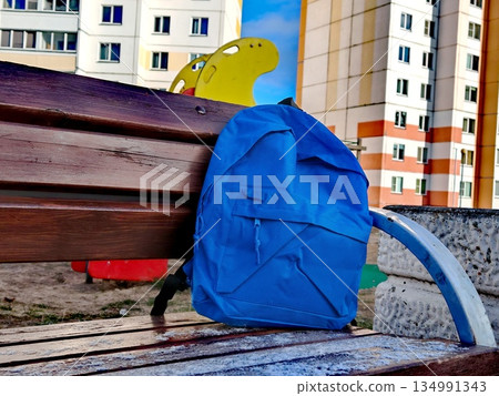 Blue Backpack Left on Bench. Blue school backpack placed on wooden park bench in urban playground area with residential buildings in background. Blue Backpack Left on Bench. Blue school backpack placed on wooden park bench in urban playground area with residential buildings in background. 134991343