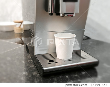 Espresso Machine Dispensing Coffee. Automatic espresso machine preparing coffee into white paper cup placed on drip tray in modern kitchen setting. 134991345