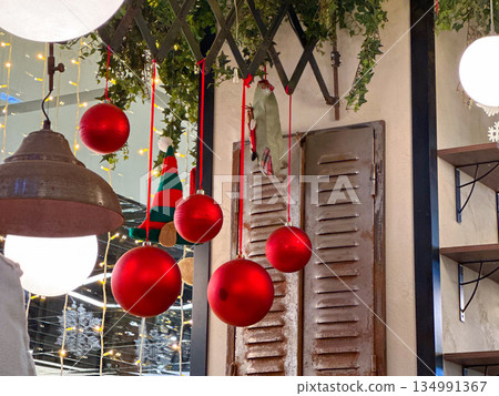 Red Christmas ornaments hanging among lights and greenery indoors. Festive interior styling, holiday decor, warm illumination, seasonal atmosphere, and cozy visual accents in public space. 134991367