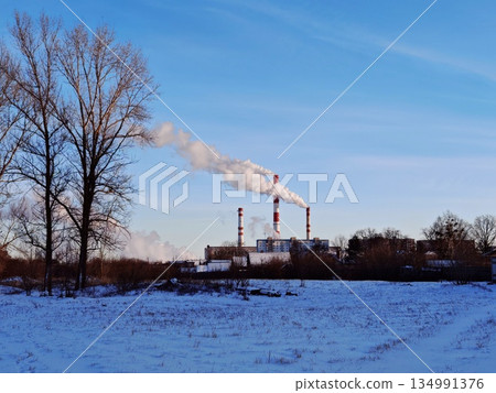 Industrial Factory with Smoking Chimneys in Winter Landscape. Factory complex emitting smoke from tall striped chimneys against clear sky with snowy field and bare trees in foreground. 134991376