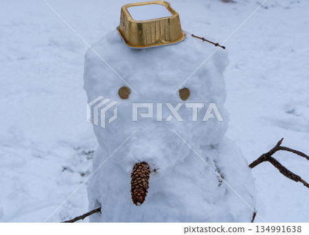 Macro view of a snowman's face made from white snow. Features include eyes made of small objects, a nose made from a pine cone, and a gold-colored plastic container as a hat. Blurred snow background. Macro view of a snowman's face made from white snow. Features include eyes made of small objects, a nose made from a pine cone, and a gold-colored plastic container as a hat. Blurred snow background. 134991638