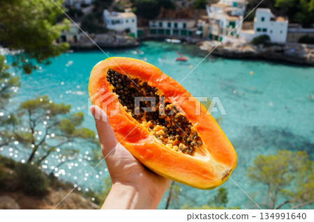 Half of fresh ripe papaya cut open with seeds in woman hand with the background of shining turquoise sea bay in summer sunny day Half of fresh ripe papaya cut open with seeds in woman hand with the background of shining turquoise sea bay in summer sunny day 134991640