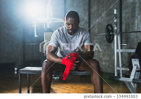 Fitness workout in gym. African American man fighter preparing for fight wrapping hands with red boxing wraps sports protective bandages in gym. Strong man ready for fight boxing sparring training Fitness workout in gym. African American man fighter preparing for fight wrapping hands with red boxing wraps sports protective bandages in gym. Strong man ready for fight boxing sparring training 134991883