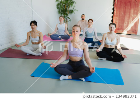 Yoga mindfulness meditation. Group of sporty people doing exercise in studio for class for yoga wellness mindfulness meditation. Men women practicing yoga sitting in lotus pose meditating relaxing Yoga mindfulness meditation. Group of sporty people doing exercise in studio for class for yoga wellness mindfulness meditation. Men women practicing yoga sitting in lotus pose meditating relaxing 134992371
