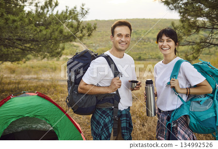 Happy tourist couple with backpacks by tent, sharing thermos at forest sunset Happy tourist couple with backpacks by tent, sharing thermos at forest sunset 134992526