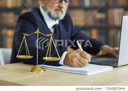 Attorney writing notes at desk with laptop and justice scales in law library Attorney writing notes at desk with laptop and justice scales in law library 134992944