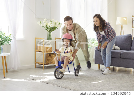 Happy parents help daughter ride tricycle indoors, playful family moment at home Happy parents help daughter ride tricycle indoors, playful family moment at home 134992945