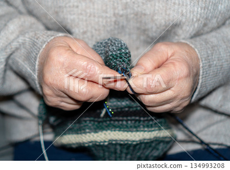 A detailed macro shot of a senior woman's hands knitting a garment using dark green melange wool yarn and metallic needles. The focus is on the intricate movement of the fingers and the texture of the A detailed macro shot of a senior woman's hands knitting a garment using dark green melange wool yarn and metallic needles. The focus is on the intricate movement of the fingers and the texture of the 134993208