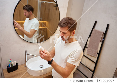 Man standing in bathroom reading label on cosmetic bottle near sink 134994097