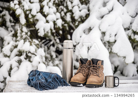 Outdoor winter still life with brown hiking boots, stainless steel thermos and metal mug placed on a snowy wooden table in front of snow covered trees, symbolizing warmth, adventure and cold season 134994265