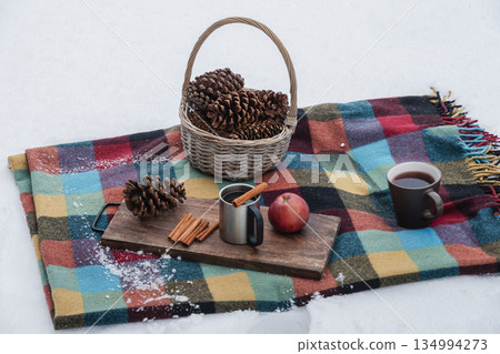 Colorful winter picnic scene with checkered blanket on snow, basket of pine cones, apple, cinnamon and stainless mug with hot drink, symbolizing outdoor winter leisure and warmth. 134994273