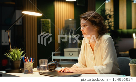 Overburdened woman prepares to leave office by turning off the lamp, doing overtime late at night due to intense workload and pressure. Uninterested discouraged worker. Camera B. Overburdened woman prepares to leave office by turning off the lamp, doing overtime late at night due to intense workload and pressure. Uninterested discouraged worker. Camera B. 134995034