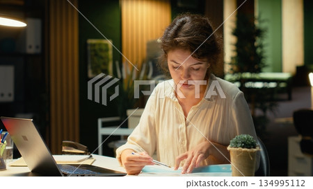 Manager researching numbers and finance figures in a dark office after hours, documenting updates and organizing the agenda for business presentation. Setting targets at night. Camera B. 134995112