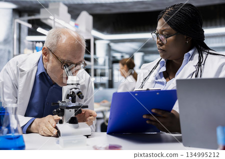 Scientist helped by lab assistant to examine bacterial cultures using microscope. Elderly man and colleague in laboratory ensure consumer safety, checking for microscopic pathogens with optical device 134995212