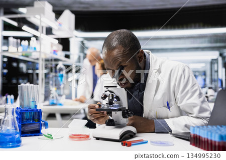 Materials scientist in lab checks product surface for tiny defects using microscope. African american man in research facility ensures manufacturing standards, examines small parts with optical device 134995230