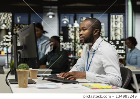 Black young analyst focused on IT company development with digital tools, review analytics and performance metrics at workstation. Staff planning solutions and aligning objectives. 134995249