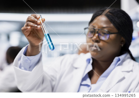 Closeup of chemical engineer in lab inspecting blue liquid clarity in test tube against light. Research facility worker verifying chemical purity standards for industrial product manufacturing process 134995265