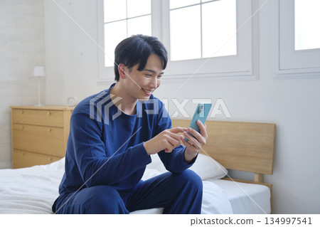 A man in his twenties sitting on a bed and using a smartphone A man in his twenties sitting on a bed and using a smartphone 134997541