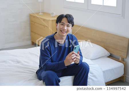 A man in his 20s sitting on a bed and holding a smartphone, looking at the camera, portrait 134997542