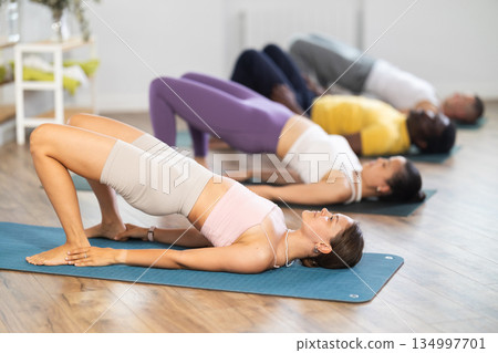 Young woman coach and students perform chatush padasana during training in studio Young woman coach and students perform chatush padasana during training in studio 134997701