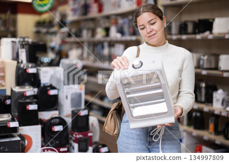 Positive young woman choosing portable heater in hypermarket Positive young woman choosing portable heater in hypermarket 134997809