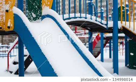 Snow covered blue playground slide with yellow climbing panels in winter park with residential buildings background during cold season Snow covered blue playground slide with yellow climbing panels in winter park with residential buildings background during cold season 134998014