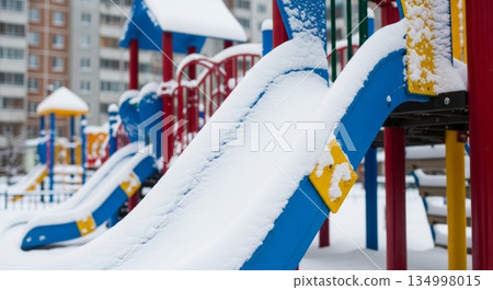 Snow covered colorful children playground equipment with blue slides and yellow accents in winter urban residential area with apartment buildings 134998015