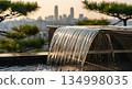 Urban waterfall fountain feature cascading into pool with blurred city skyline and acacia trees during golden hour sunset lighting 134998035