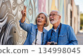 Senior european couple exploring street art mural wall wearing matching denim shirts while sightseeing with vintage camera around neck pointing at colorful urban artwork 134998045