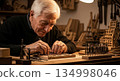 Senior European craftsman with gray hair working on detailed wood inlay project using precision carving tools in traditional workshop with warm lighting 134998046