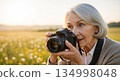 Senior european woman with silver bob haircut taking photographs with black DSLR camera in golden sunset meadow field with wildflowers 134998048