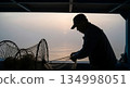 Silhouetted fisherman repairing fishing nets on boat deck during misty sunrise over calm ocean waters 134998051