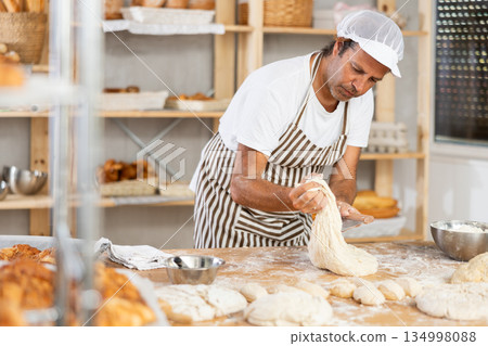 Professional male baker uses knife to cut raw dough to make croissants or buns Professional male baker uses knife to cut raw dough to make croissants or buns 134998088