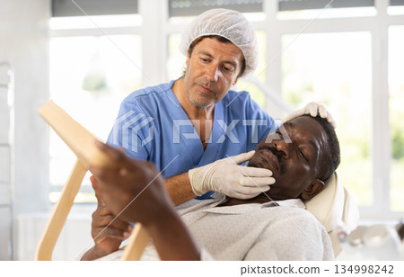 Cosmetologist showing results of facial procedure to dark-skinned man 134998242