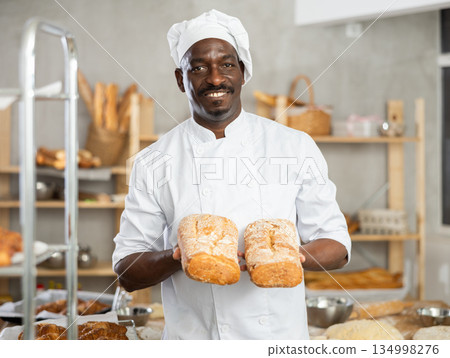 Portrait of experienced baker with tasty hot bread and baguettes in hands in kitchen Portrait of experienced baker with tasty hot bread and baguettes in hands in kitchen 134998276