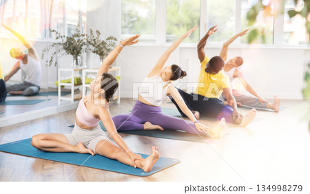 Young woman coach and students perform Janu Shirshasana during training in studio Young woman coach and students perform Janu Shirshasana during training in studio 134998279