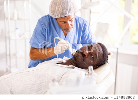 Male specialist uses ultrasonic import nozzle to care African man client face skin 134998908