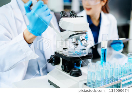 Closeup of Asian scientists experimenting with blue liquids and microscopes in a modern lab, representing biotechnology, chemistry, and medical innovation. 135000674