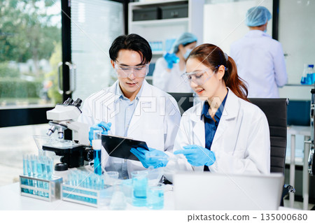 Scientists collaborating in a modern lab with microscopes and blue liquids, representing biotechnology, chemistry, and medical research innovation. 135000680
