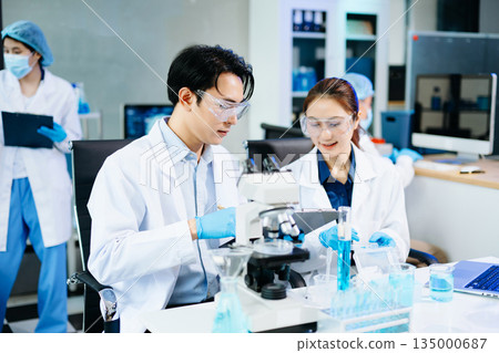 Scientists collaborating in a modern lab with microscopes and blue liquids, representing biotechnology, chemistry, and medical research innovation. 135000687