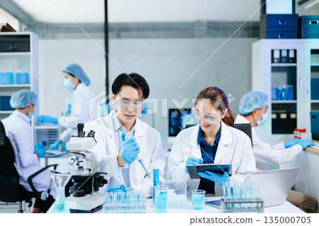 Scientists collaborating in a modern lab with microscopes and blue liquids, representing biotechnology, chemistry, and medical research innovation. 135000705