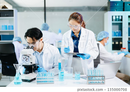 Scientists collaborating in a modern lab with microscopes and blue liquids, representing biotechnology, chemistry, and medical research innovation. 135000708