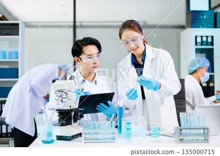 Scientists collaborating in a bright modern lab with blue liquids and microscopes, symbolizing teamwork, biotechnology, chemistry, and medical innovation. 135000715