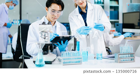 Scientists collaborating in a bright modern lab with blue liquids and microscopes, symbolizing teamwork, biotechnology, chemistry, and medical innovation. 135000716
